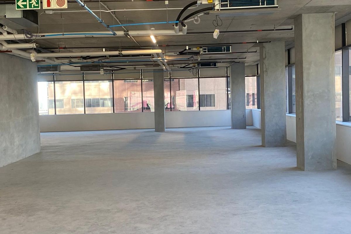 An open-plan office space featuring an industrial aesthetic with exposed ceiling infrastructure, concrete columns, and large windows providing ample natural light.