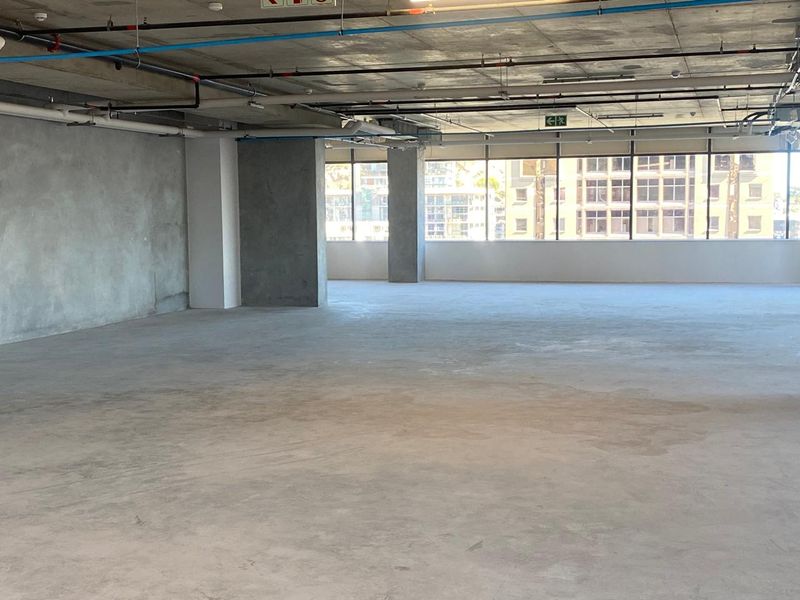 A spacious, open-plan office area featuring an industrial aesthetic with exposed concrete ceilings, visible utility services, and large windows providing ample natural light.