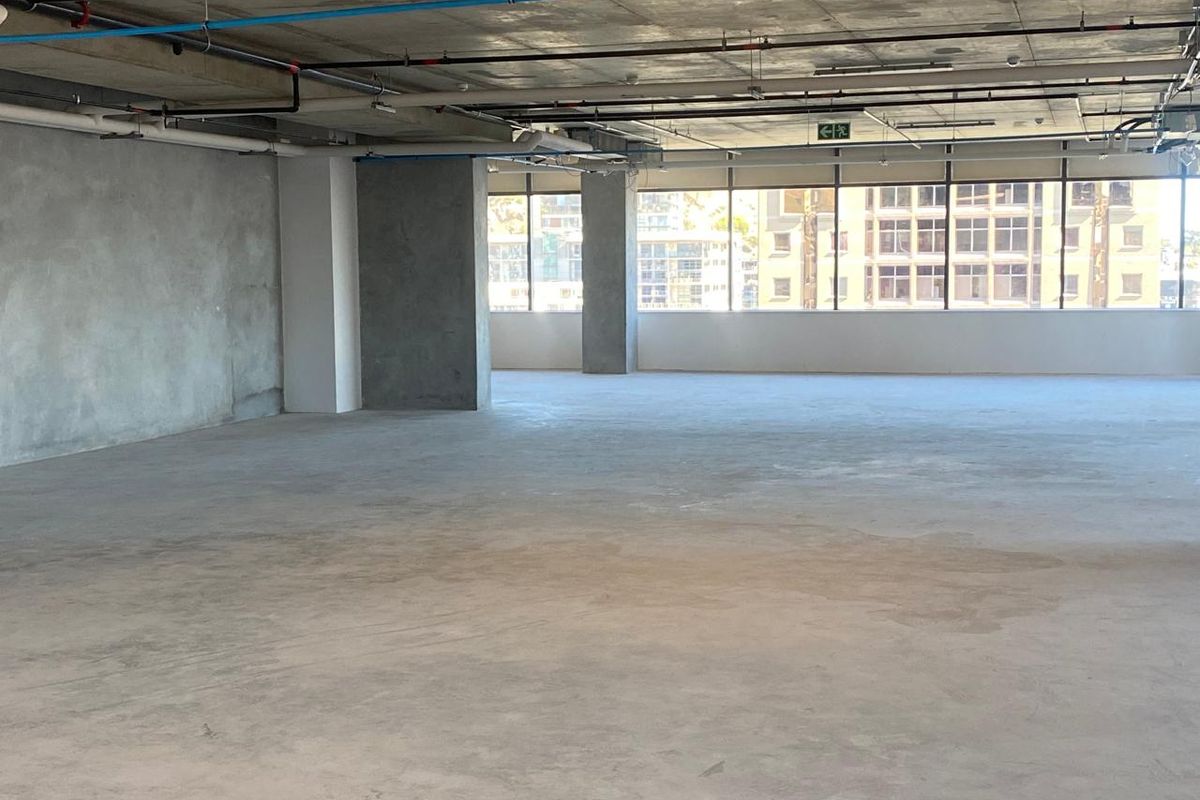 A spacious, open-plan office area featuring an industrial aesthetic with exposed concrete ceilings, visible utility services, and large windows providing ample natural light.