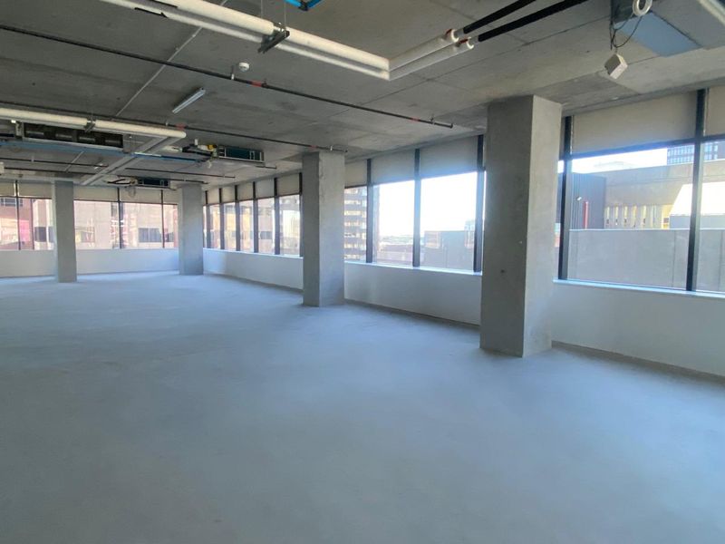 An open-plan office space featuring an industrial aesthetic with exposed concrete ceilings, large windows, and polished concrete flooring.