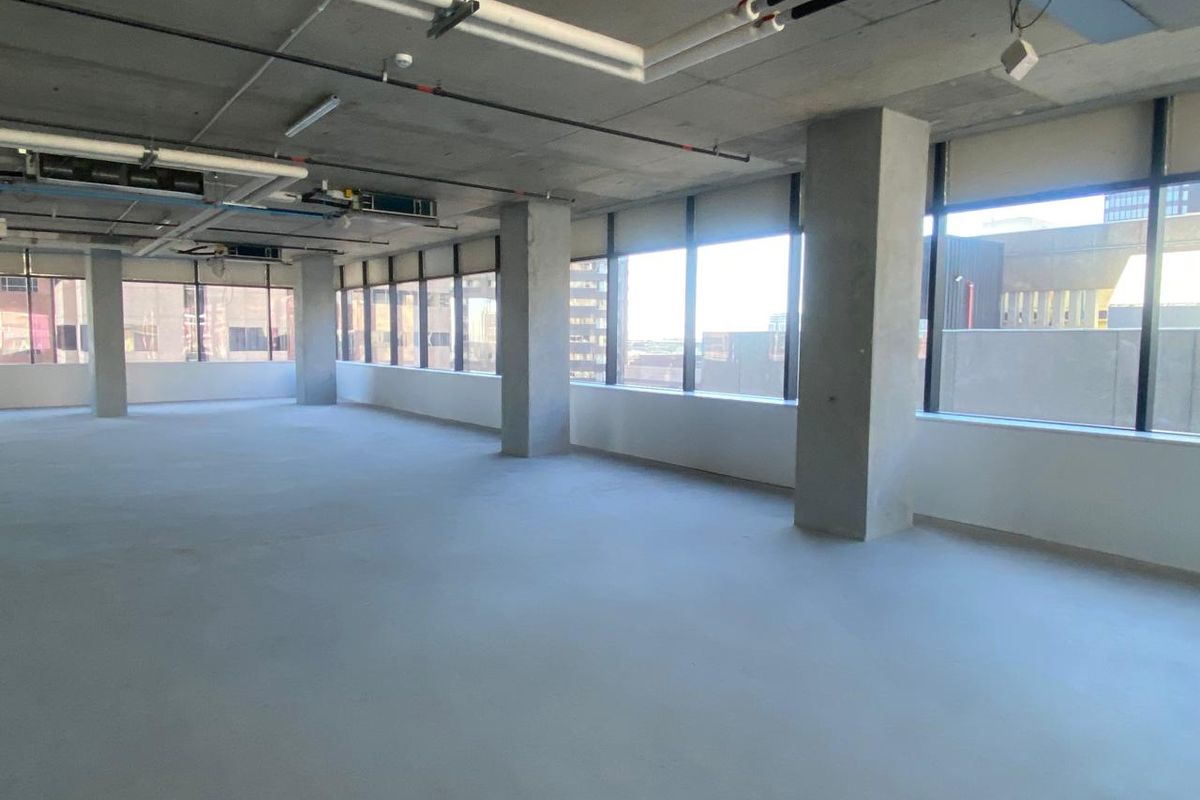 An open-plan office space featuring an industrial aesthetic with exposed concrete ceilings, large windows, and polished concrete flooring.