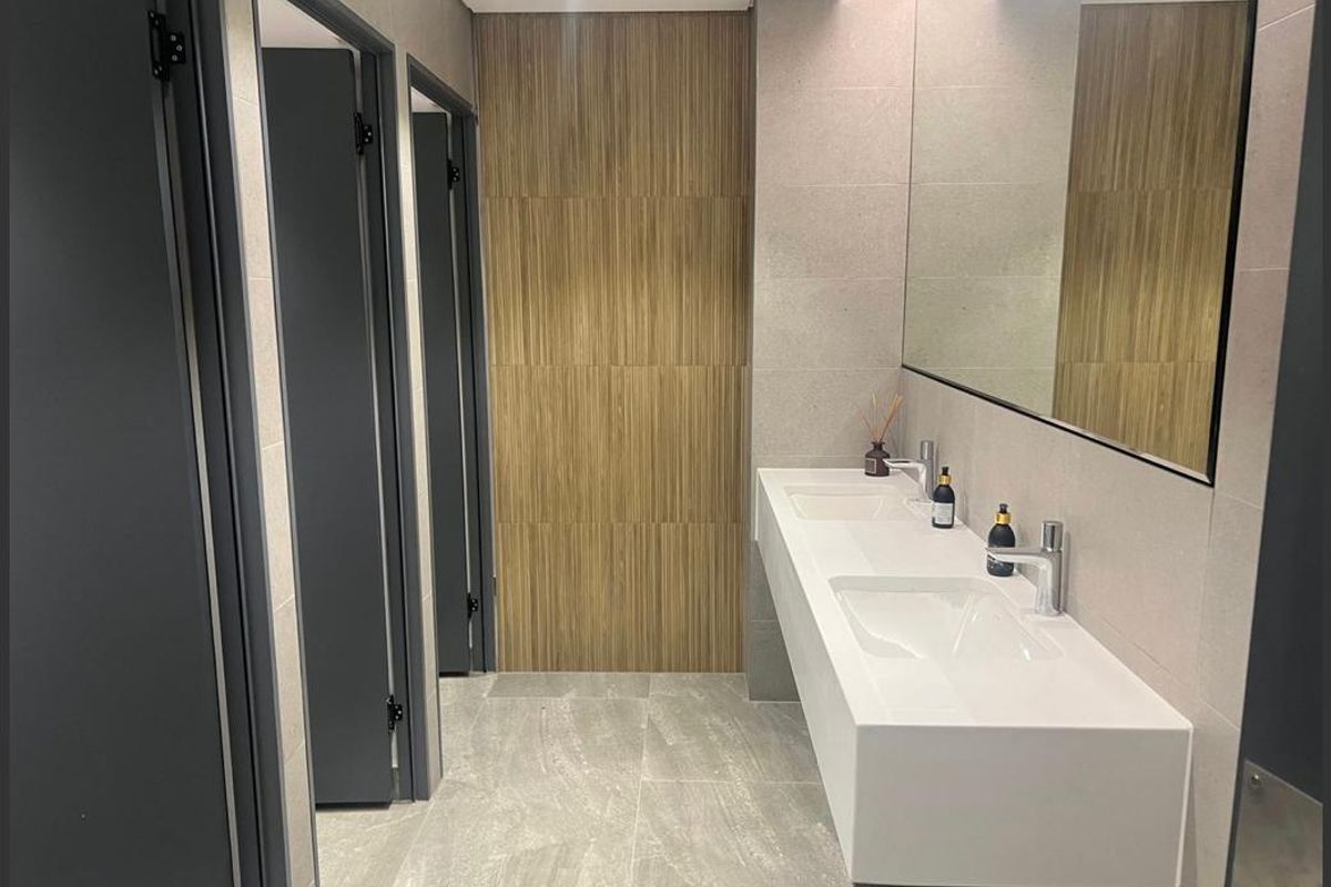 Modern office restroom featuring a sleek double vanity with integrated sinks, large wall-mounted mirror, and contemporary wood-look tile accent wall.