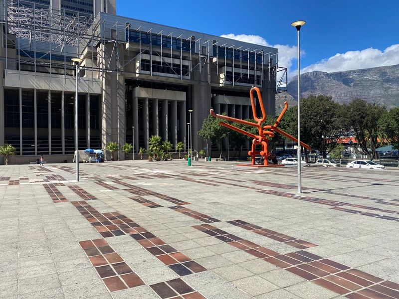 A wide-angle view of a paved commercial plaza featuring modern architectural design, street lighting, and a prominent outdoor sculpture with mountain views in the background.