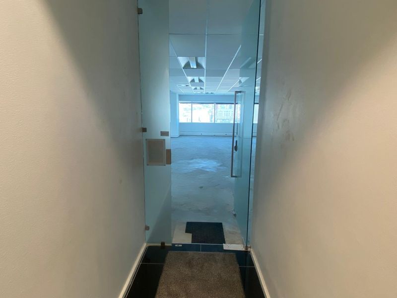 An empty office space featuring a glass entrance door, suspended ceiling with recessed lighting, and large windows providing natural light.