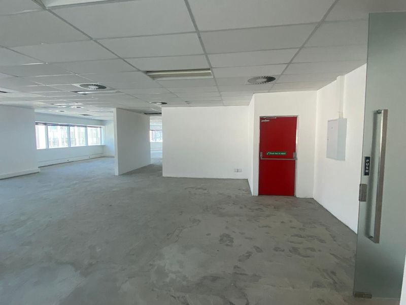 An open-plan office space featuring a suspended ceiling, recessed lighting, and a prominent red fire door, currently presented as a blank canvas.