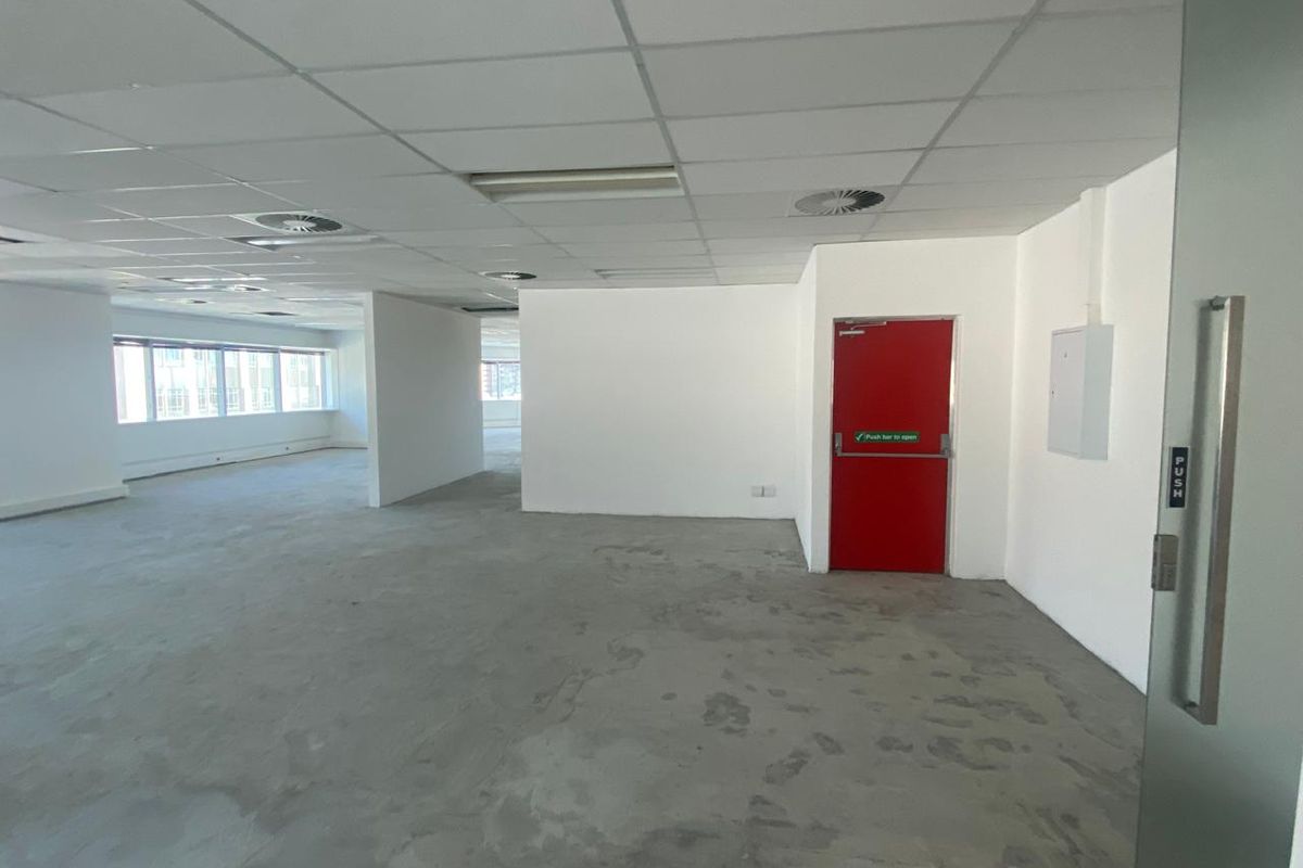 An open-plan office space featuring a suspended ceiling, recessed lighting, and a prominent red fire door, currently presented as a blank canvas.