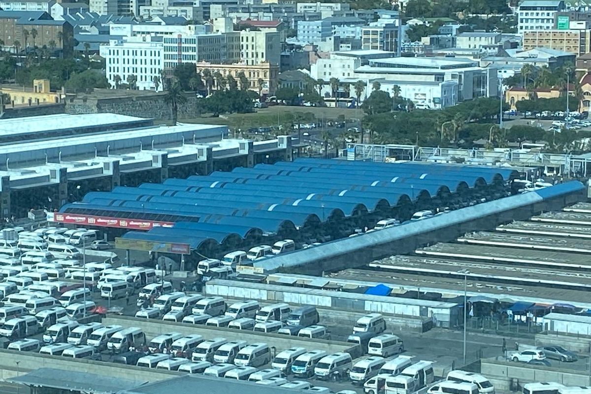 An elevated aerial view of a large transport terminal facility featuring extensive blue metal roofing and a busy parking area for commuter vehicles.
