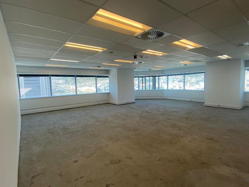 An open-plan office space featuring a suspended ceiling, recessed lighting, and large windows offering panoramic city views. The carpet flooring shows signs of wear.