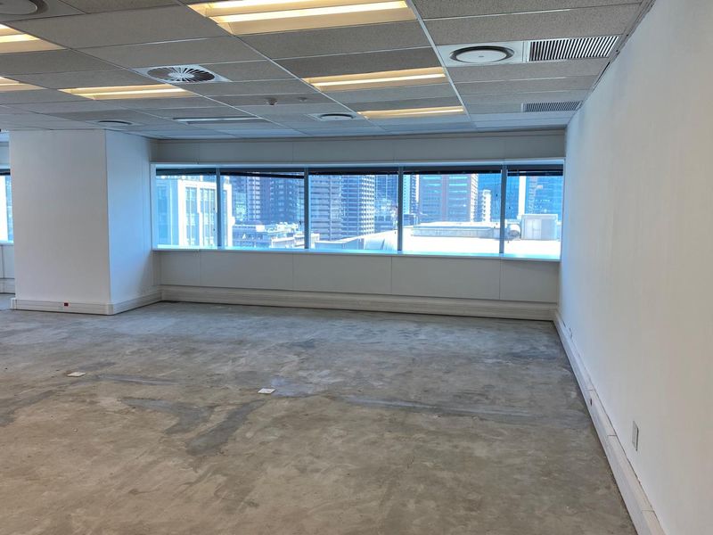 An empty, open-plan office space featuring a suspended ceiling, recessed lighting, and large windows with city views, requiring new floor finishes.