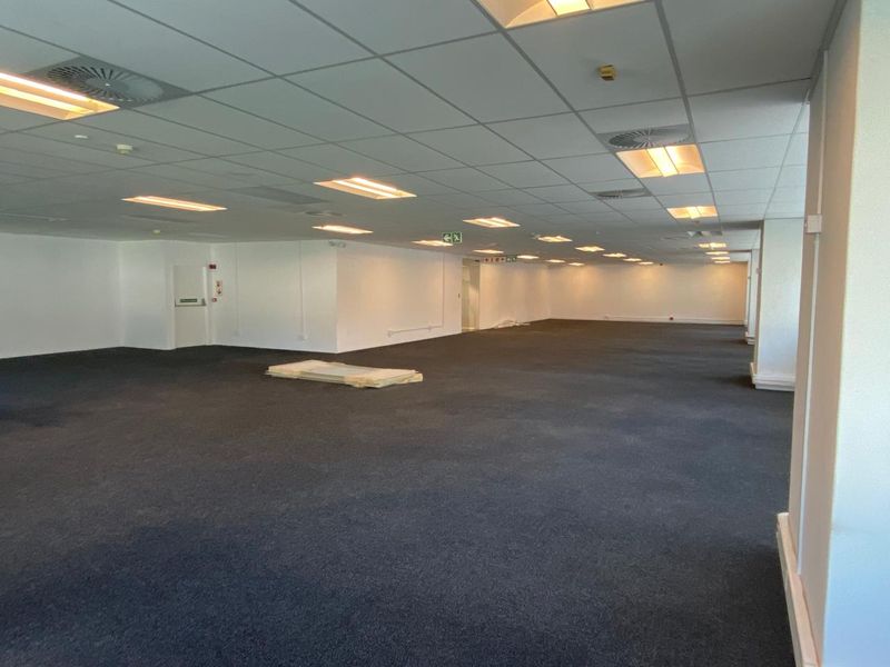 A large, open-plan office space featuring a suspended ceiling with recessed lighting, air conditioning diffusers, and dark carpeting throughout.
