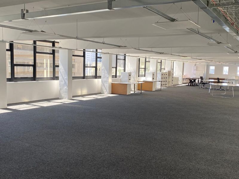 Spacious open-plan office area featuring large windows, exposed ceiling infrastructure, and neutral carpet flooring, providing a bright and professional workspace.
