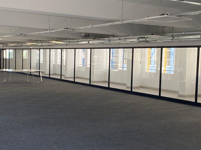 Spacious open-plan office area featuring modern glass partitioning, exposed ceiling infrastructure, and neutral carpet flooring, offering a clean and professional workspace.