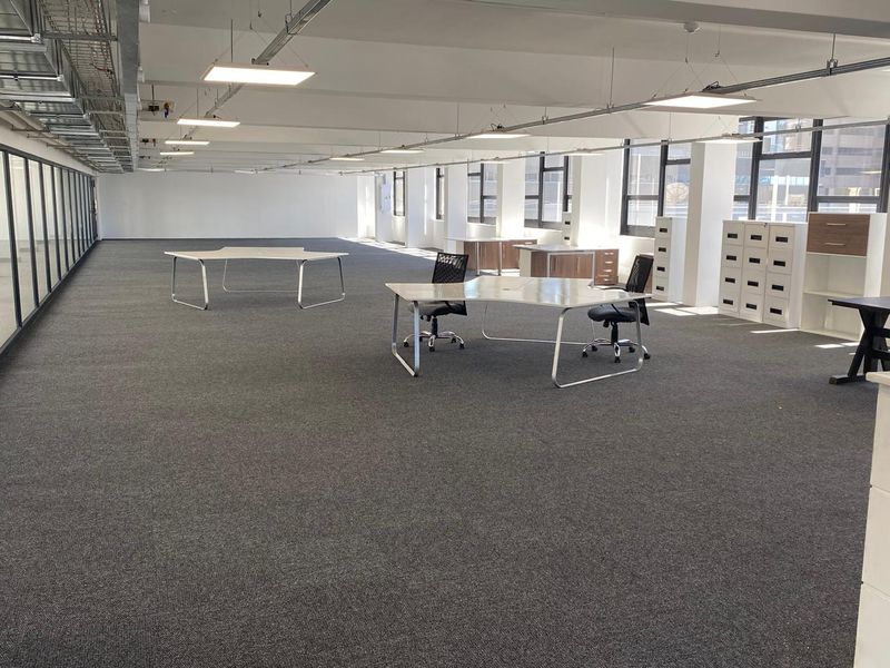Spacious open-plan office area featuring exposed ceiling ducting, modern LED panel lighting, and large windows providing ample natural light.