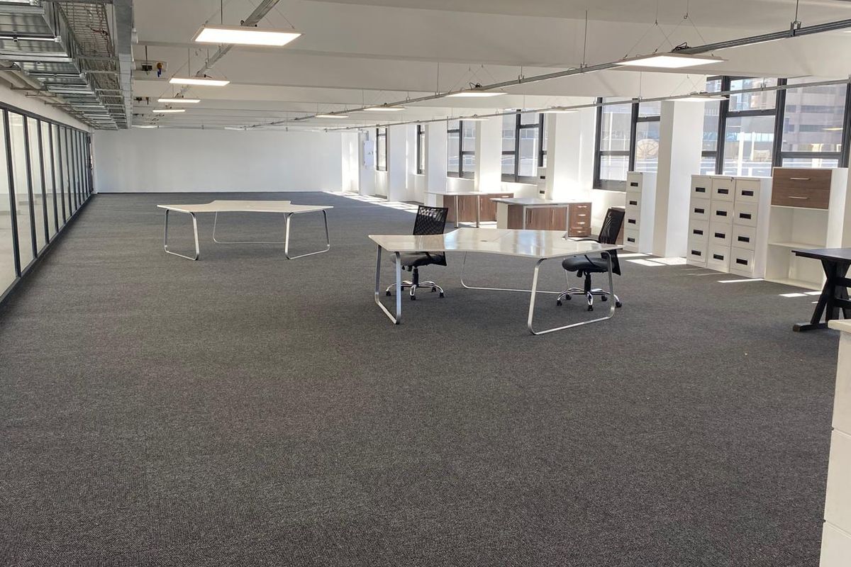 Spacious open-plan office area featuring exposed ceiling ducting, modern LED panel lighting, and large windows providing ample natural light.