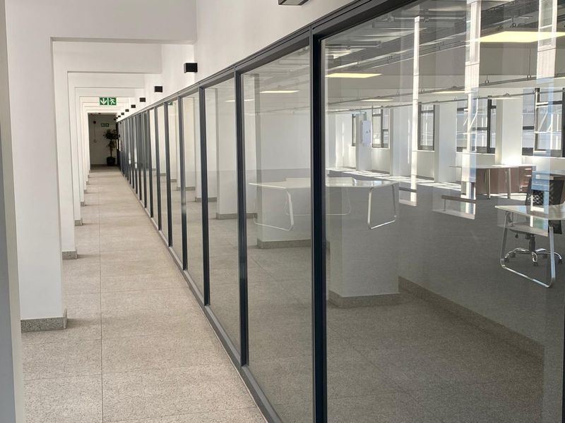 A bright, modern office hallway featuring extensive glass partitioning, clean tiled floors, and professional recessed lighting, creating an open and airy workspace environment.