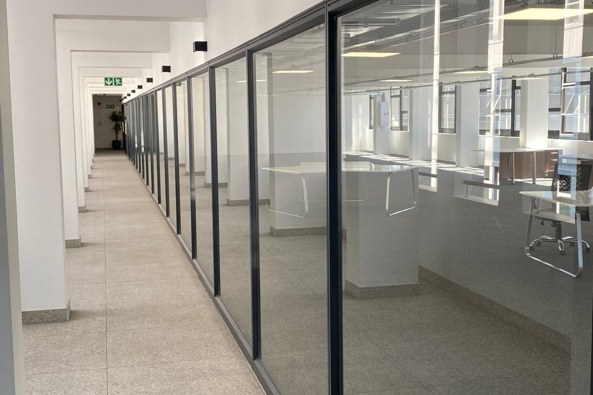 A bright, modern office hallway featuring extensive glass partitioning, clean tiled floors, and professional recessed lighting, creating an open and airy workspace environment.