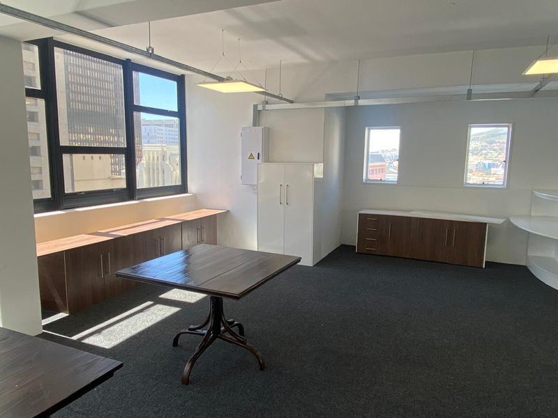 A bright, professional office space featuring built-in storage cabinetry, carpeted flooring, and large windows providing natural light and city views.