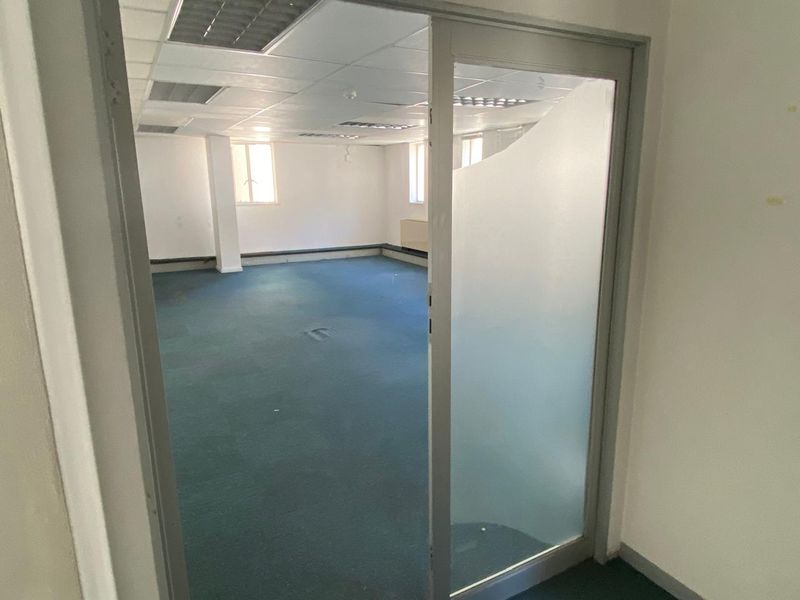 An empty commercial office space featuring a suspended ceiling, recessed lighting, and carpeted flooring, accessed via a frosted glass door.