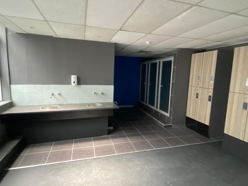 A commercial changing room area featuring built-in lockers, dual vanity sinks, tiled flooring, and a suspended ceiling.
