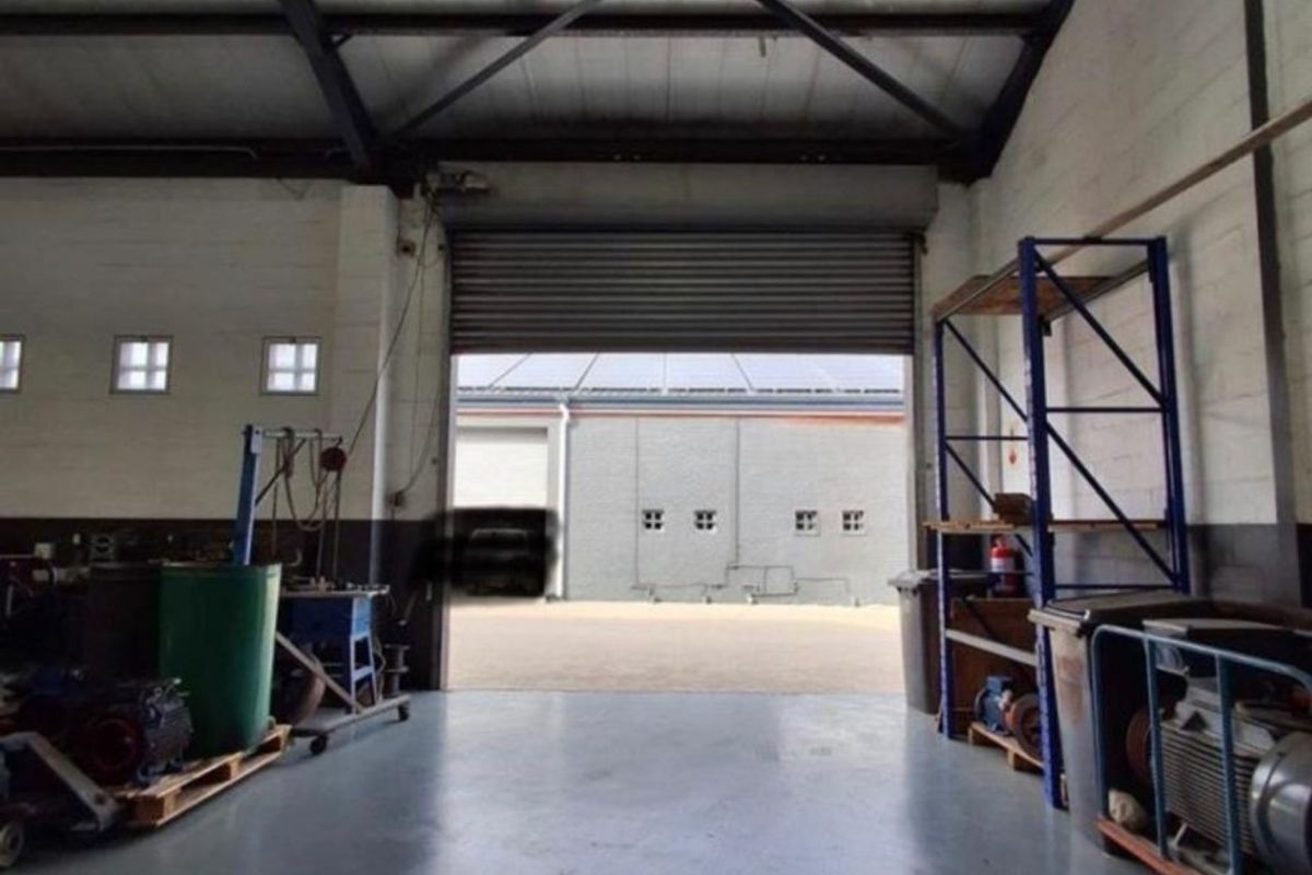 Industrial warehouse space featuring high ceilings with exposed steel trusses, concrete flooring, and a large roller shutter door for vehicle or equipment access.
