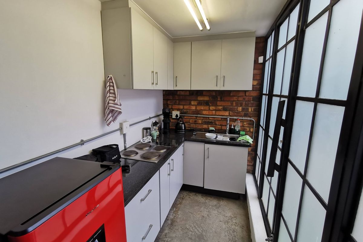 Compact kitchenette featuring white cabinetry, dark countertops, a built-in electric hob, and a stainless steel sink, set against a rustic exposed brick wall.