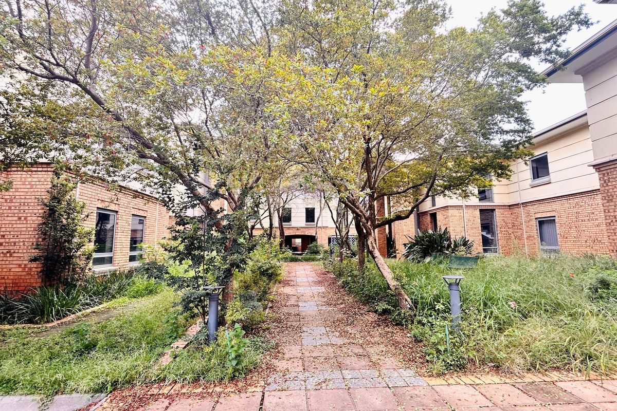 A landscaped courtyard area featuring a central paved walkway flanked by mature trees, garden beds, and brick office building facades.