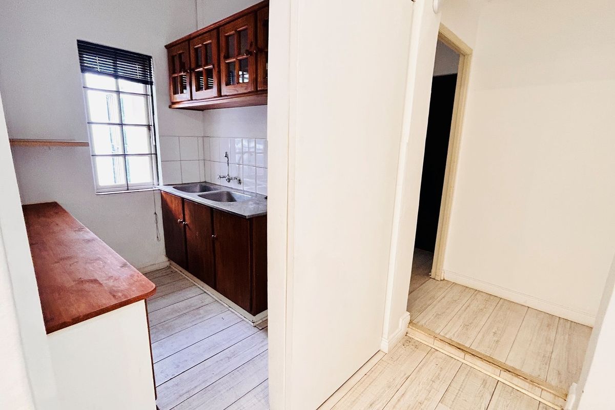 A functional kitchenette area featuring wooden cabinetry, a double sink, and tiled splashback, adjacent to a hallway with light-toned flooring.