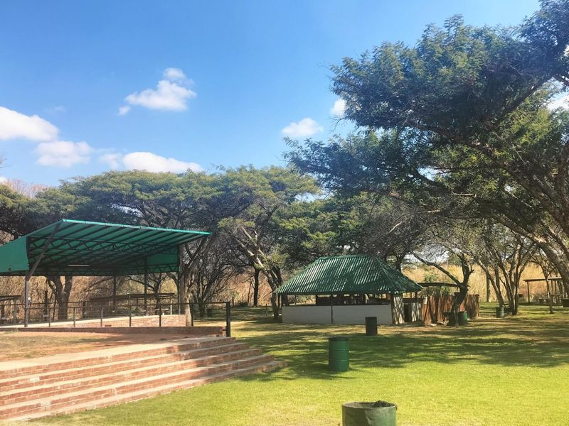Outdoor recreational area featuring a covered stage, a small service structure, and landscaped grounds with mature trees.