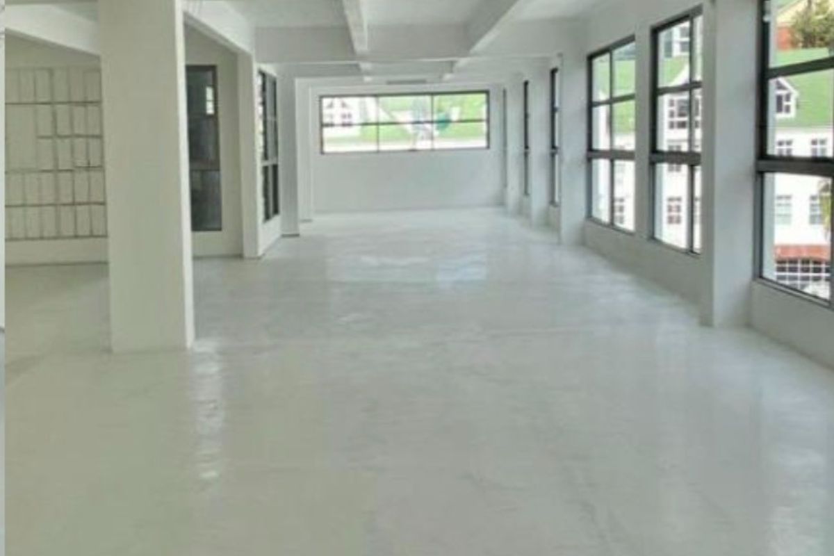 A spacious, open-plan commercial office area featuring polished concrete floors, exposed ceiling beams, and large windows providing ample natural light.