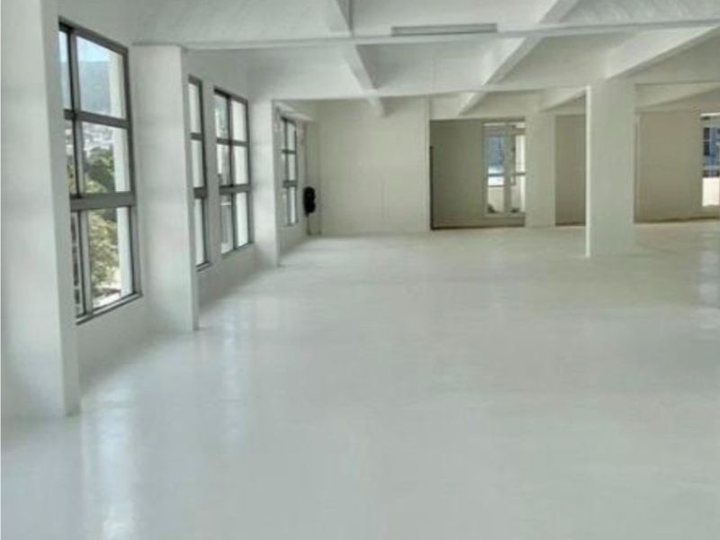 Spacious open-plan commercial office area featuring high ceilings with exposed beams, large windows providing natural light, and clean, polished flooring.
