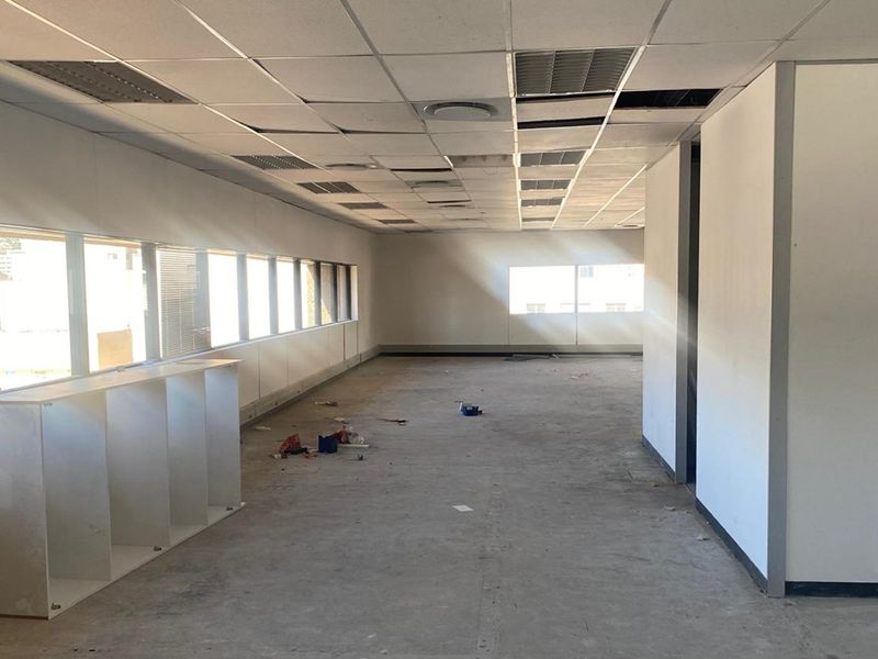 An empty, open-plan office space featuring large windows, a suspended ceiling with missing tiles, and unfinished flooring, ready for renovation.