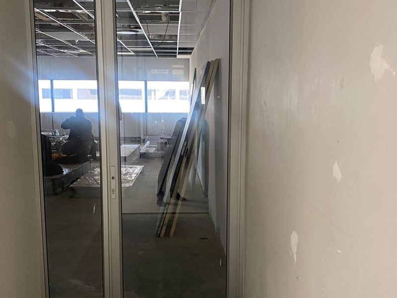 An office space currently under renovation, featuring glass partition doors, an exposed suspended ceiling grid, and unfinished wall surfaces.