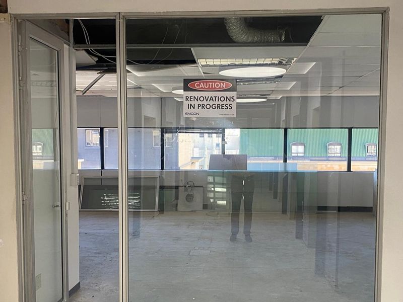 An empty office space currently undergoing renovations, featuring glass partitions, exposed ceiling ductwork, and large windows providing natural light.