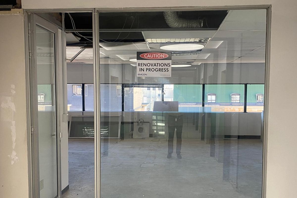 An empty office space currently undergoing renovations, featuring glass partitions, exposed ceiling ductwork, and large windows providing natural light.