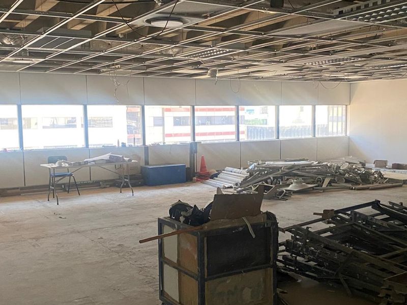 An open-plan office space currently undergoing renovation, featuring a suspended ceiling grid, large windows, and unfinished interior surfaces.