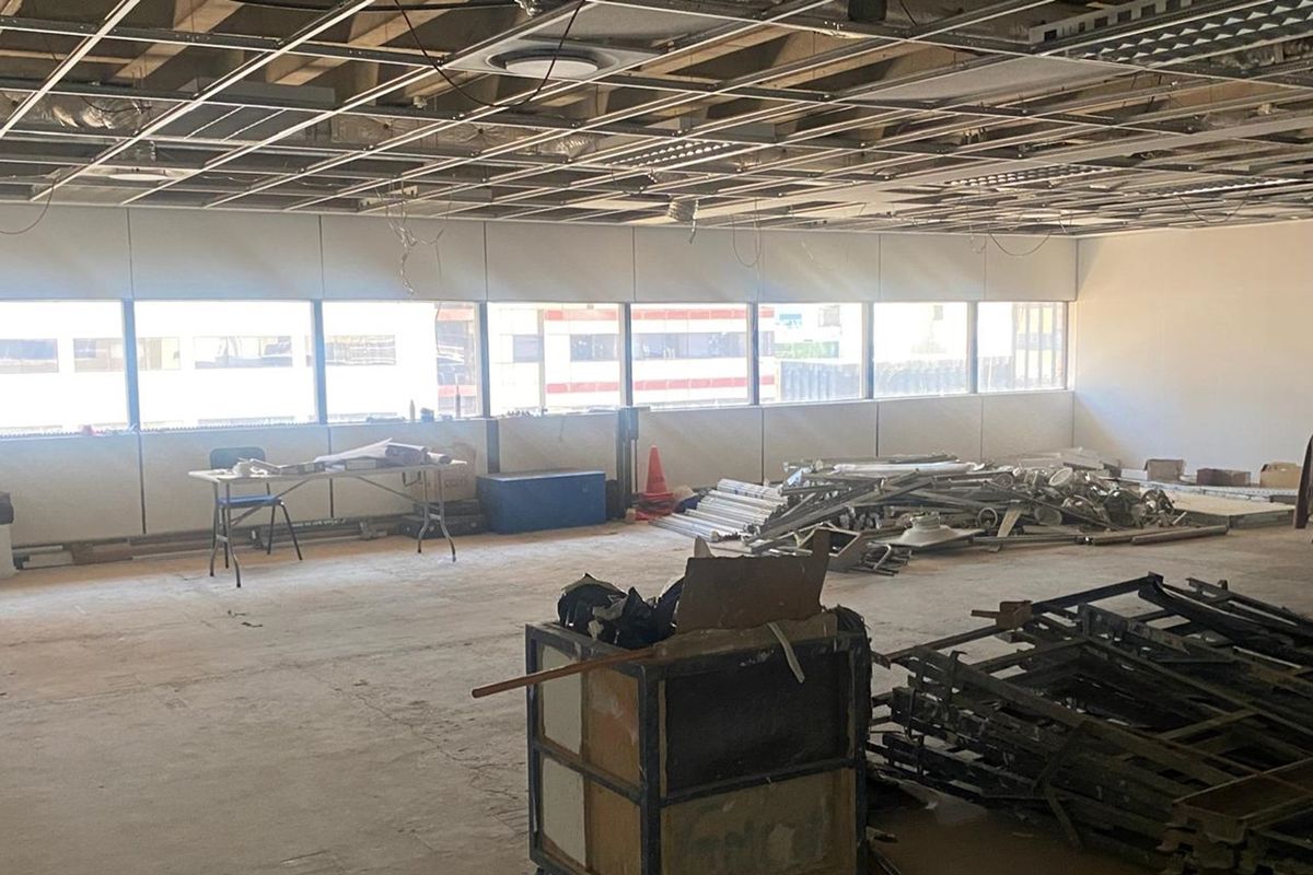 An open-plan office space currently undergoing renovation, featuring a suspended ceiling grid, large windows, and unfinished interior surfaces.