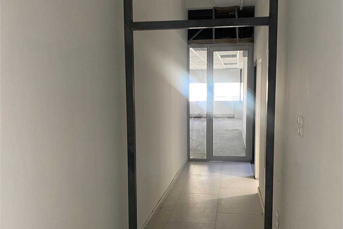 A vacant office hallway featuring a metal-framed glass door and tiled flooring, currently showing an unfinished ceiling area.