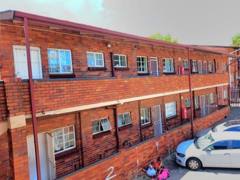 Exterior view of a two-story face brick apartment building featuring multiple casement windows, security gates, and external plumbing infrastructure.