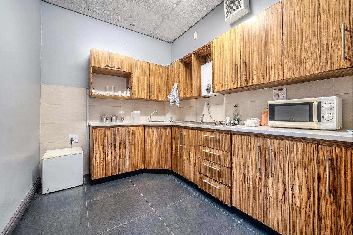 A functional kitchenette featuring wood-grain cabinetry, tiled splashbacks, a stainless steel sink, and dark floor tiles.