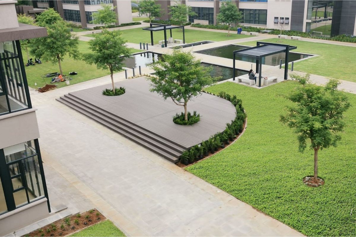 An aerial view of a modern office park courtyard featuring landscaped lawns, a raised timber deck, water features, and shaded pergola seating areas.