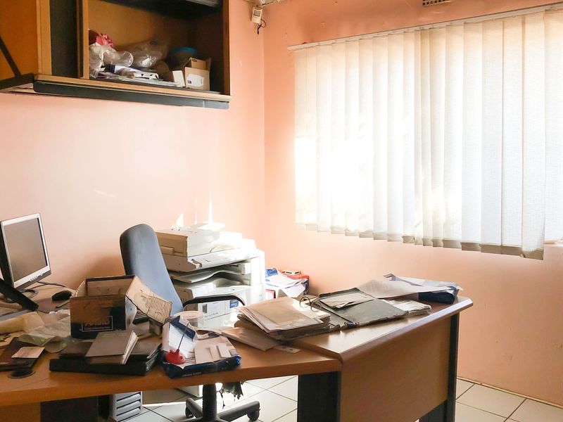 A functional office space featuring tiled flooring, vertical window blinds, and built-in wall shelving, currently used as a workspace.