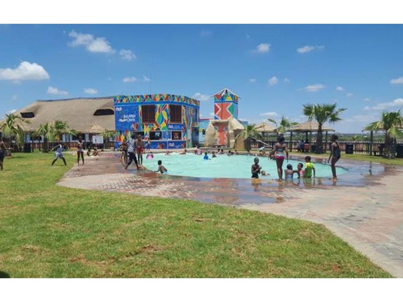 Outdoor pool area featuring a swimming pool, paved surround, landscaped lawn, and a multi-colored building structure with a thatched roof.