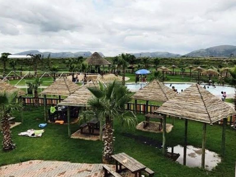 Outdoor recreational area featuring multiple thatched gazebos, landscaped lawns, and a swimming pool facility set against a scenic mountain backdrop.