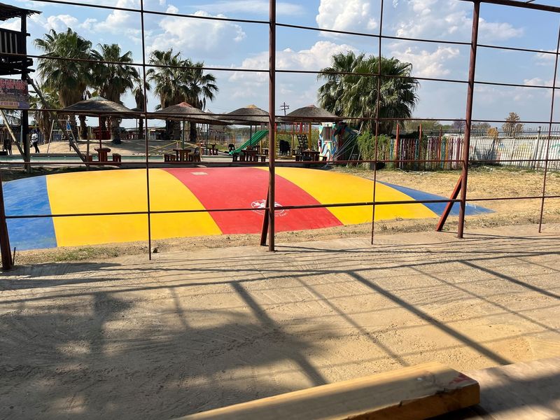 Outdoor recreational area featuring a large inflatable jumping pillow, shaded seating structures, and perimeter metal fencing.