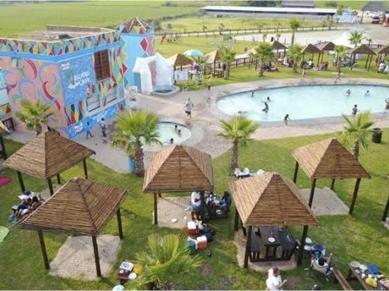 Aerial view of a recreational facility featuring a swimming pool, multiple thatched gazebos, and a brightly painted building with mural artwork.