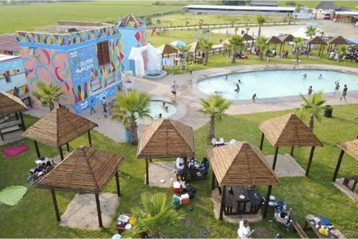 Aerial view of a recreational facility featuring a swimming pool, multiple thatched gazebos, and a brightly painted building with mural artwork.