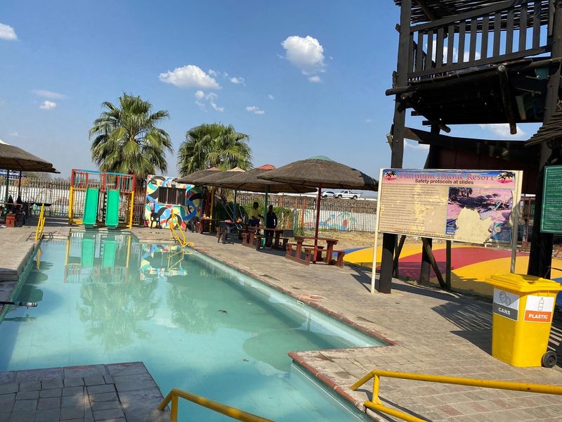 Outdoor pool area featuring a swimming pool, water slides, thatched shade structures, and a wooden observation tower within a recreational facility.