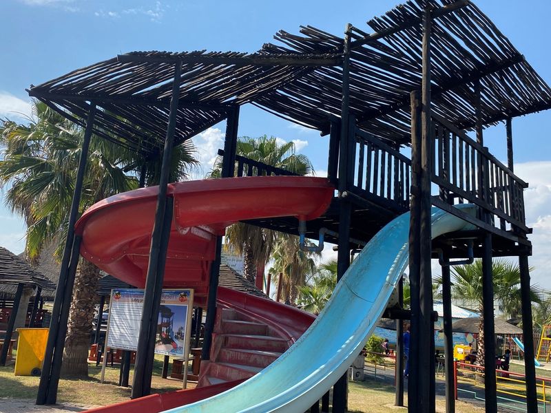 Outdoor recreational play structure featuring dual slides and a timber-framed canopy, situated within a landscaped resort or park area.