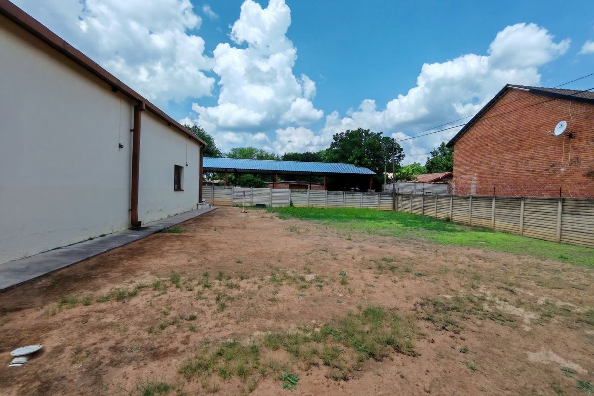 A spacious rear yard area featuring a concrete boundary wall, a covered outdoor structure, and a concrete walkway along the building side.