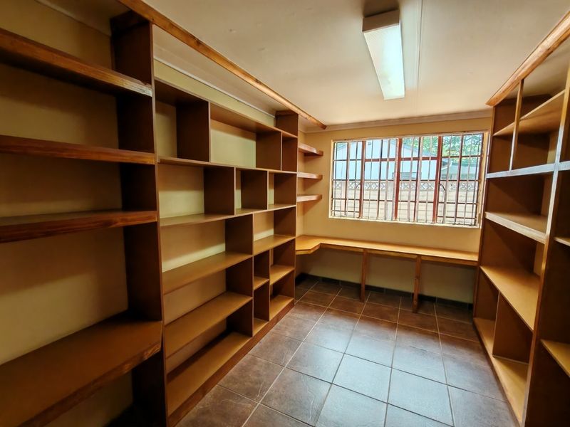 A functional office space featuring extensive built-in wooden shelving, a fixed desk, tiled flooring, and a window with security bars.